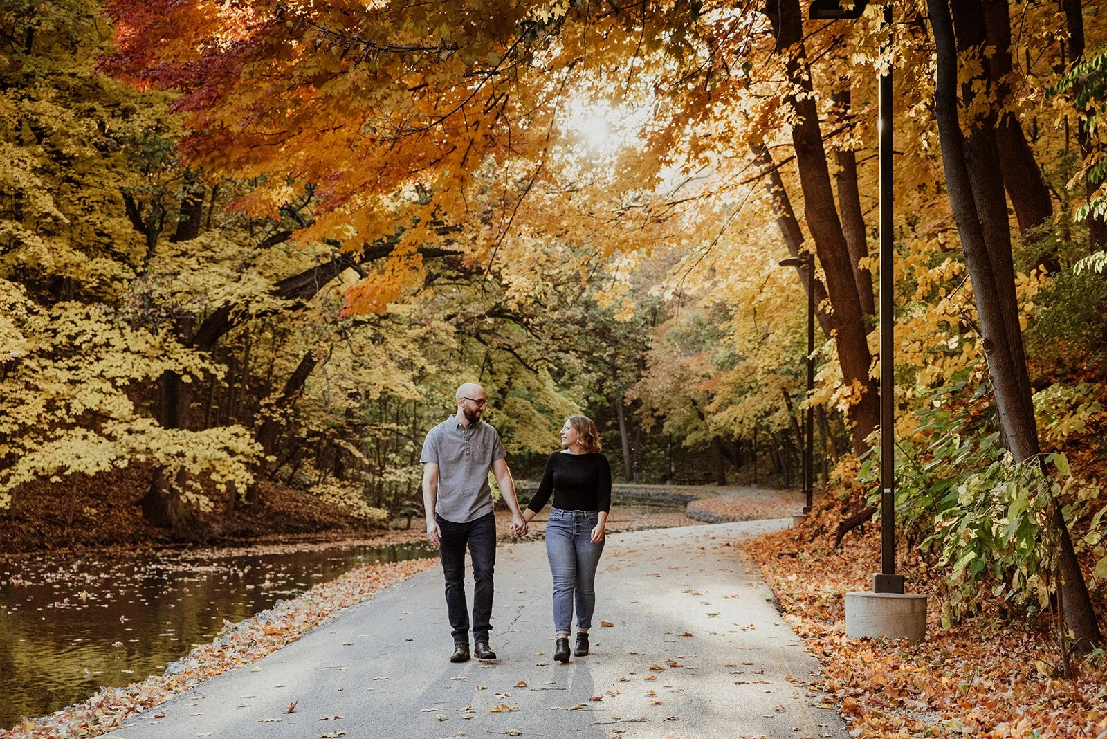 A couple walking hand in hand on a path through a park with yellow and orange autumn leaves on trees and fallen leaves on the ground, with a body of water on the left and street lamps on the right.