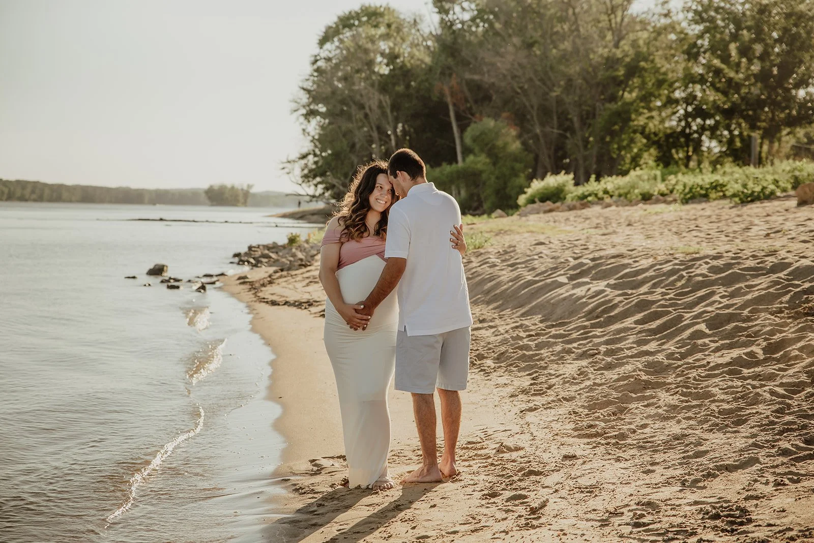 A pregnant woman and a man standing on a sandy beach by the water, touching foreheads and holding her baby bump, with trees and a calm river in the background during sunset.