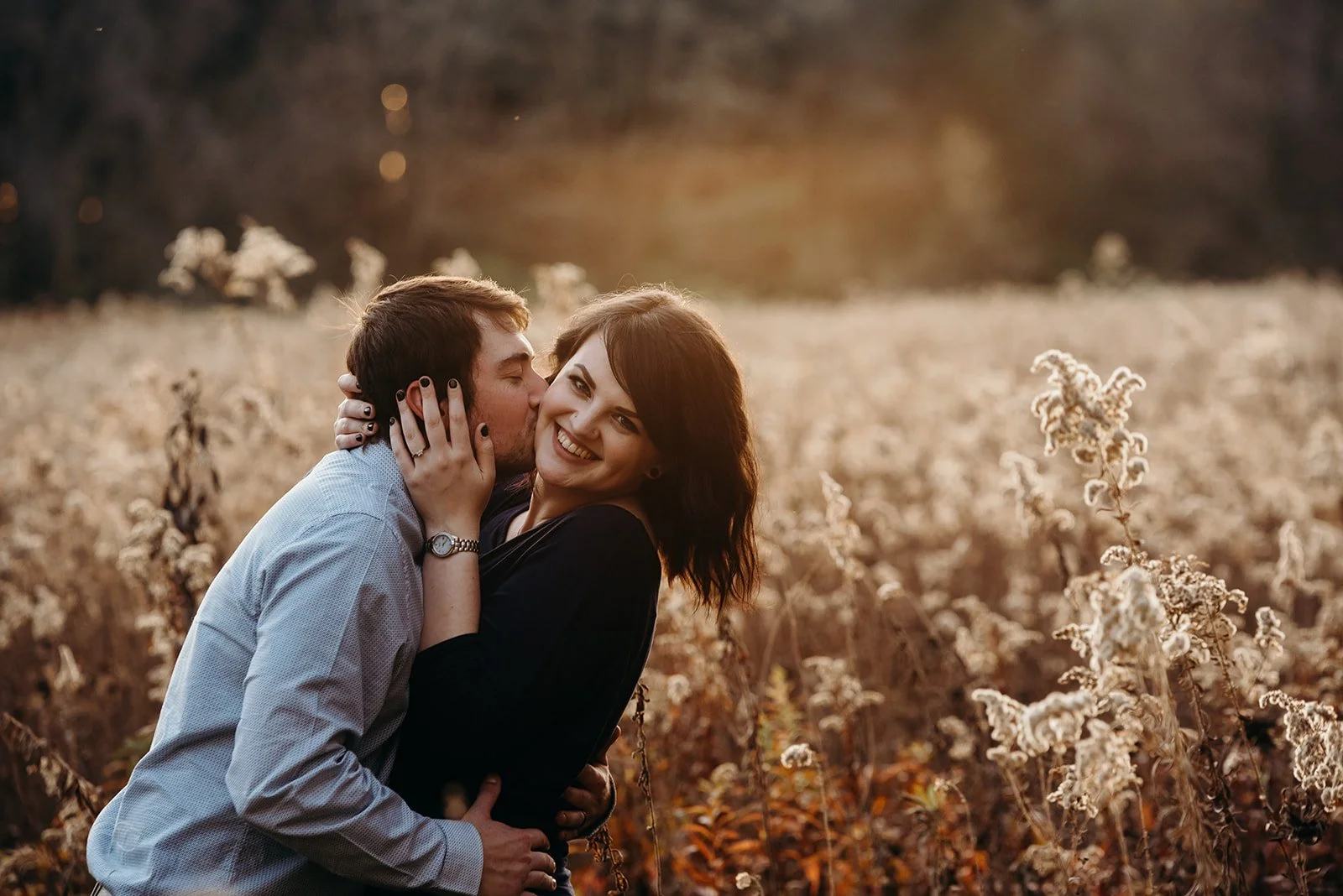 A couple embracing in a field during sunset, with the man kissing the woman's cheek and the woman smiling.