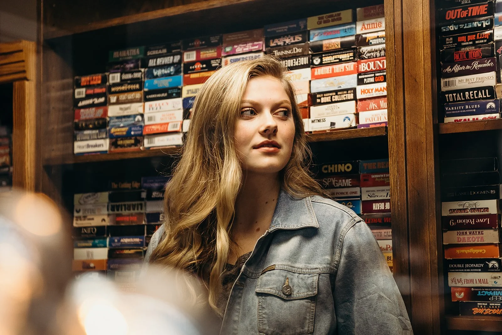 A young woman with long blonde hair in a denim jacket looking to her left in front of a bookshelf filled with multiple stacks of board games.