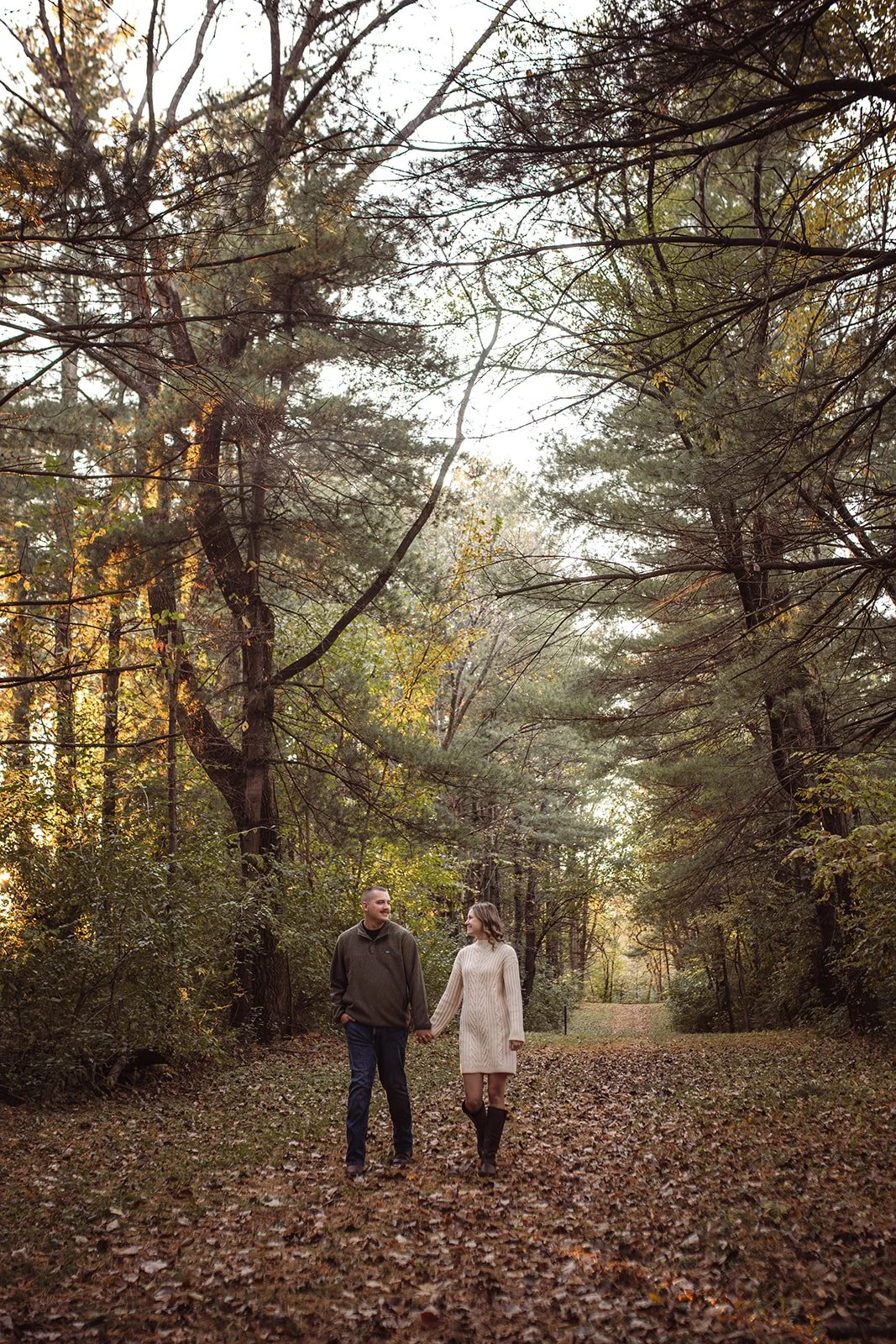 A couple walking hand-in-hand through a wooded park during fall, with trees and fallen leaves around them.