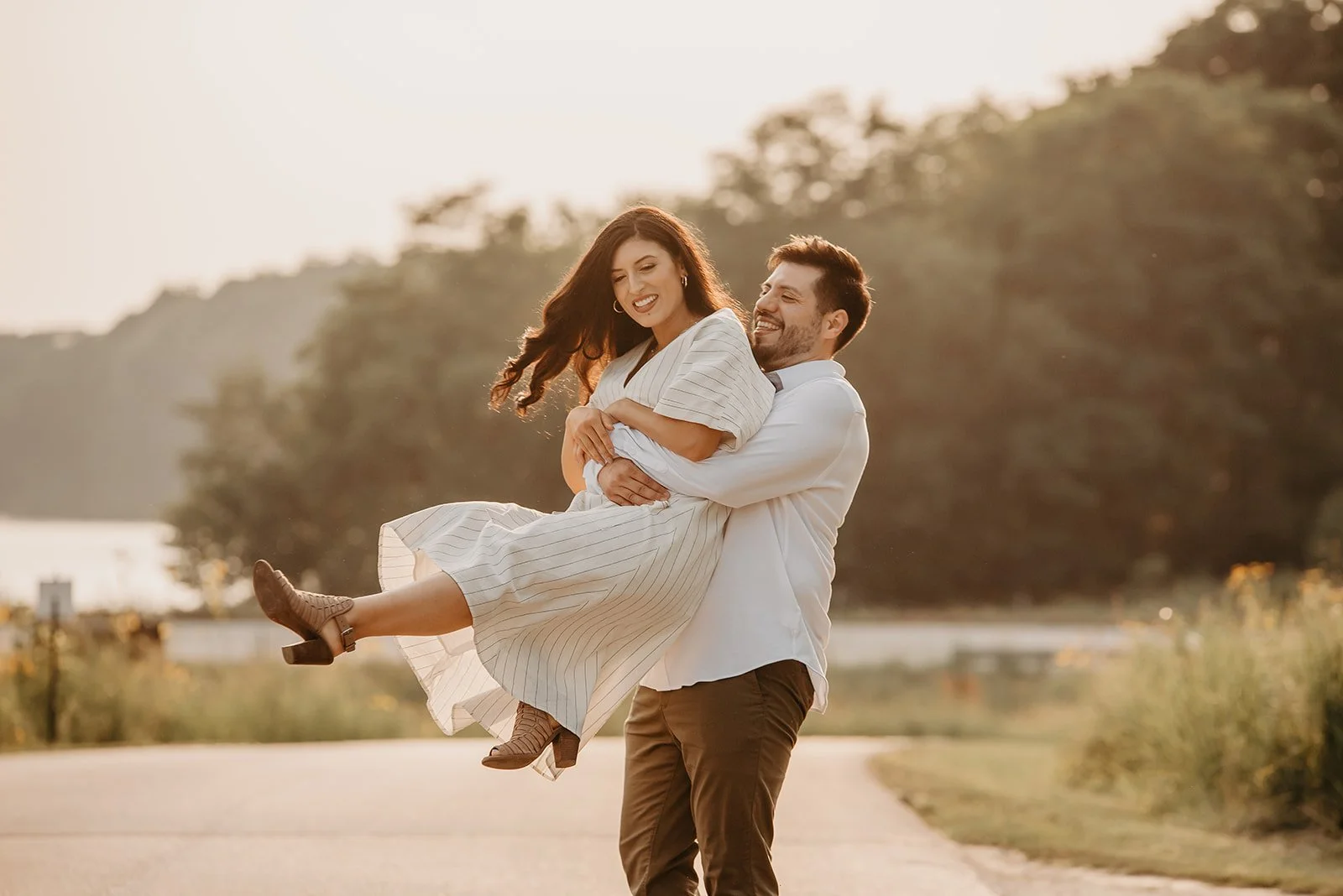 A man and woman smiling and enjoying the outdoors during sunset, with the man lifting the woman in his arms, both dressed in light-colored clothing.