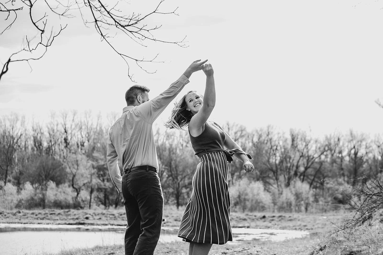 A couple dancing outdoors near a small body of water with bare trees in the background, captured in black and white.