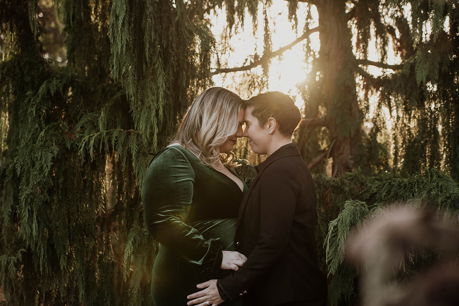 A couple standing close together, forehead to forehead, in a forest with sunlight filtering through trees.