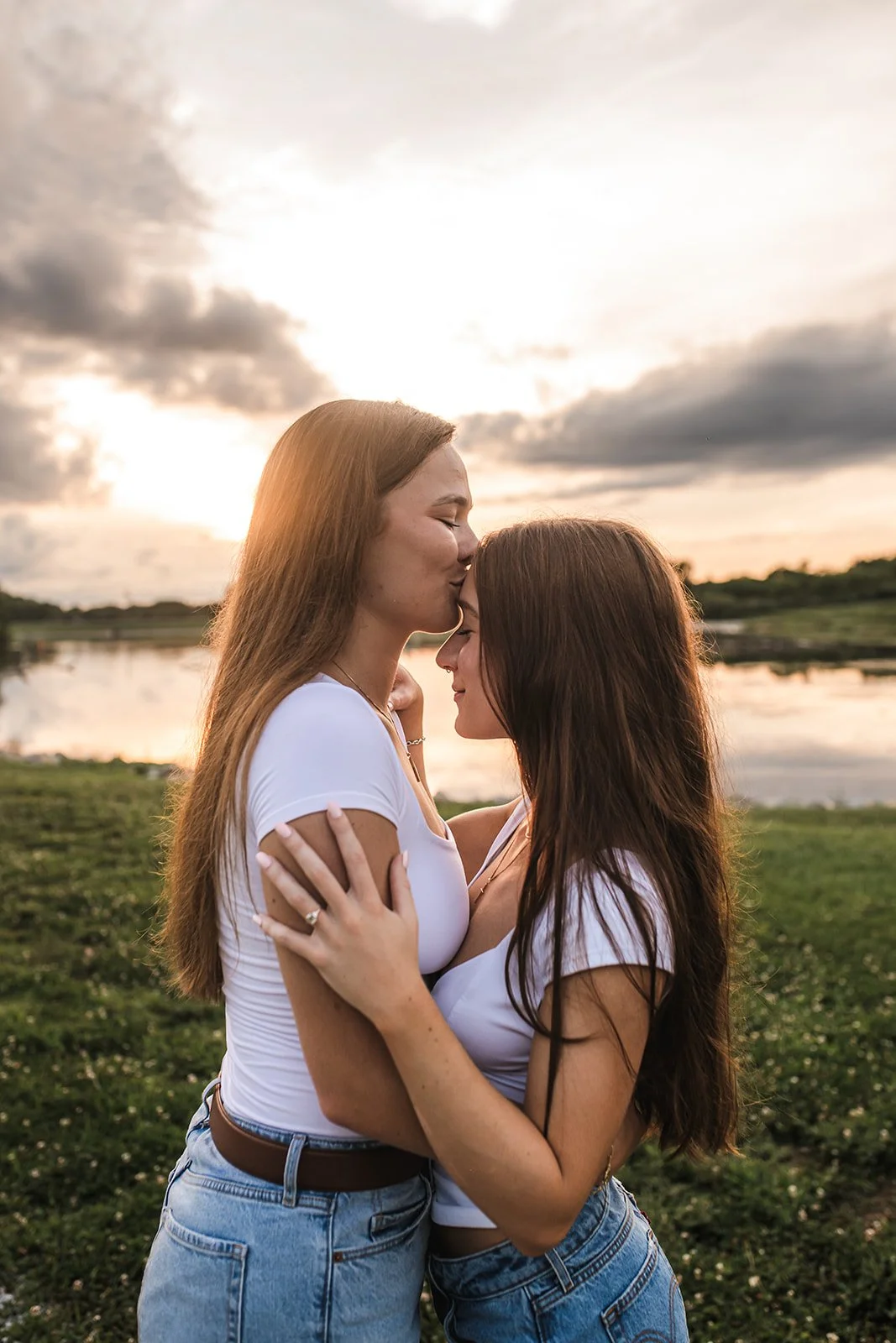 Two women embracing and sharing a kiss on the forehead outdoors at sunset by a lake.