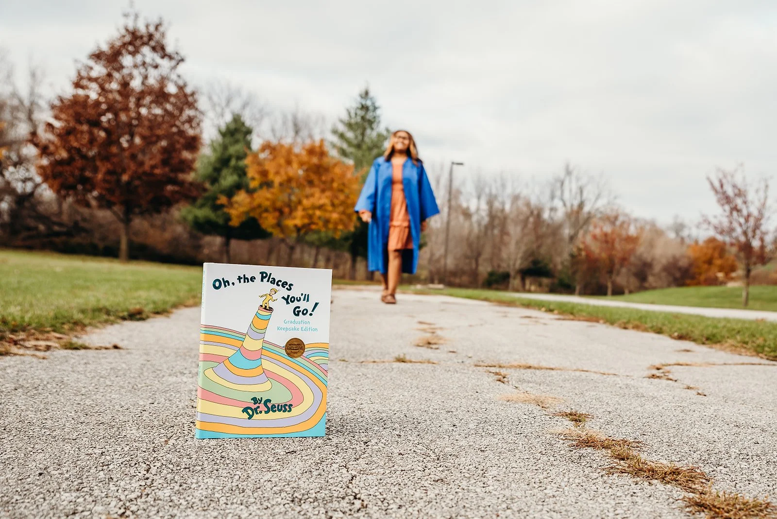 A woman in a blue graduation gown and cap walking on a park pathway with autumn trees in the background, with a copy of the book "Oh, the Places You'll Go!" by Dr. Seuss in the foreground.