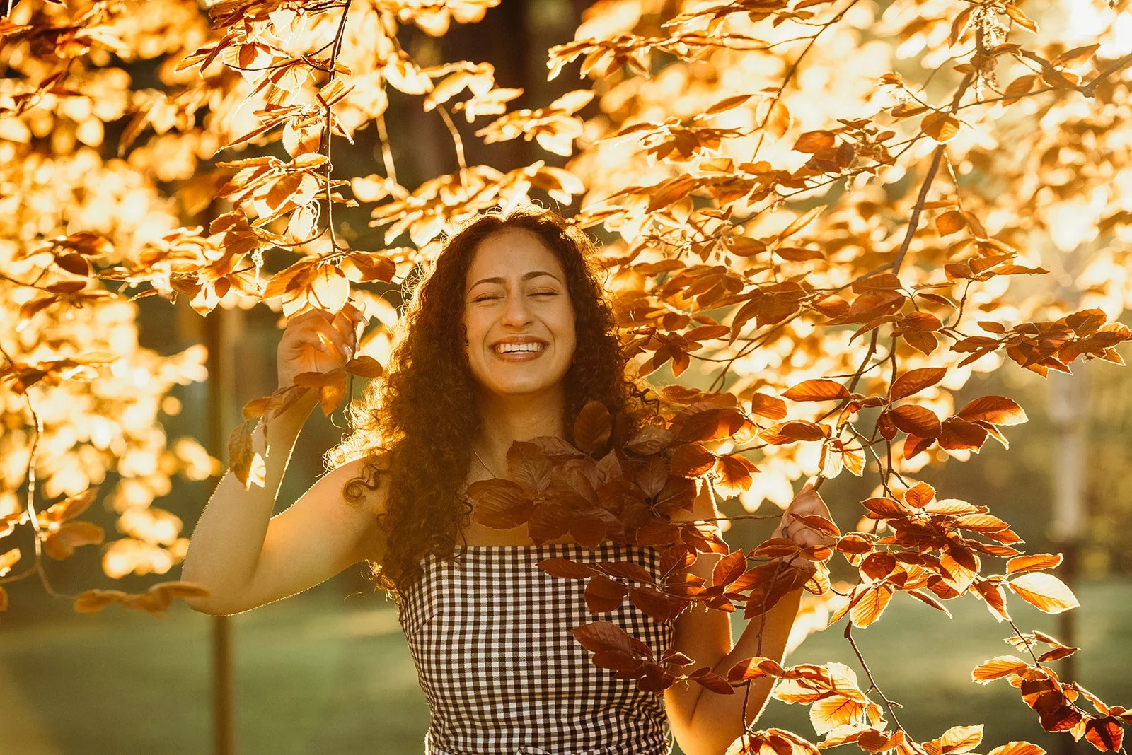 A woman with curly hair smiling and enjoying the outdoors, surrounded by orange and brown autumn leaves on a tree branch.