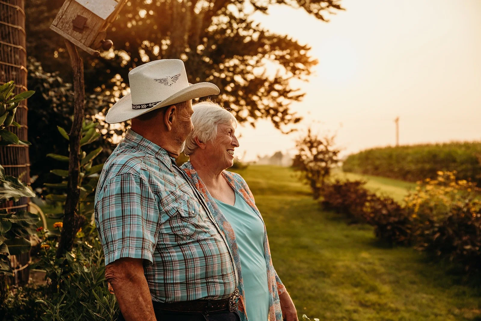 An elderly couple standing outdoors in a garden at sunset, smiling and enjoying each other's company.