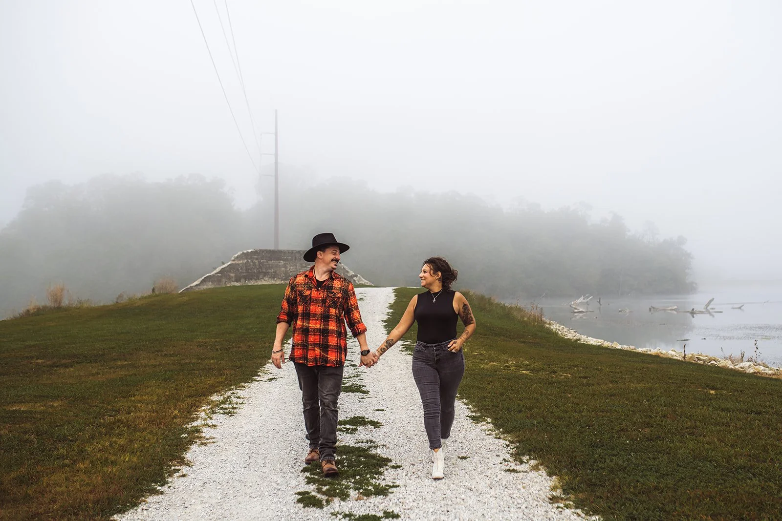 A couple holding hands and walking along a gravel path next to a body of water, with foggy trees in the background.