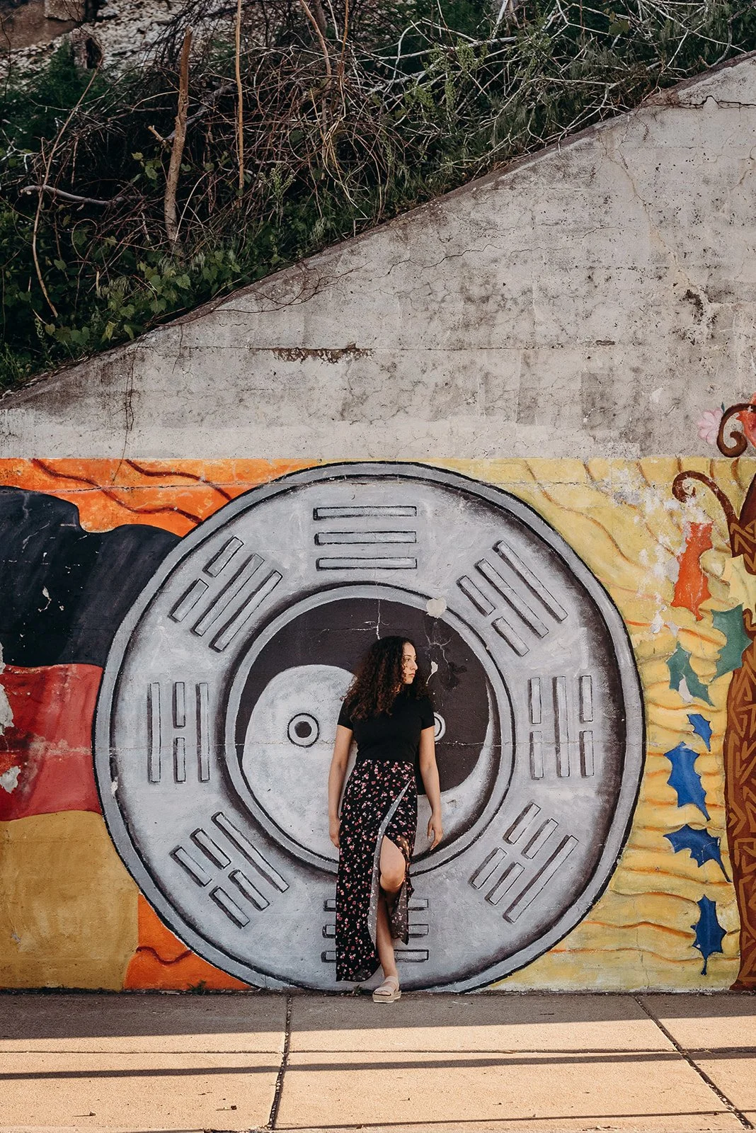 A woman standing in front of a large black, white, and gray circular mural of a traditional Chinese coin with square hole in the center, painted on a wall with colorful artistic designs and some leaves and vines at the top.
