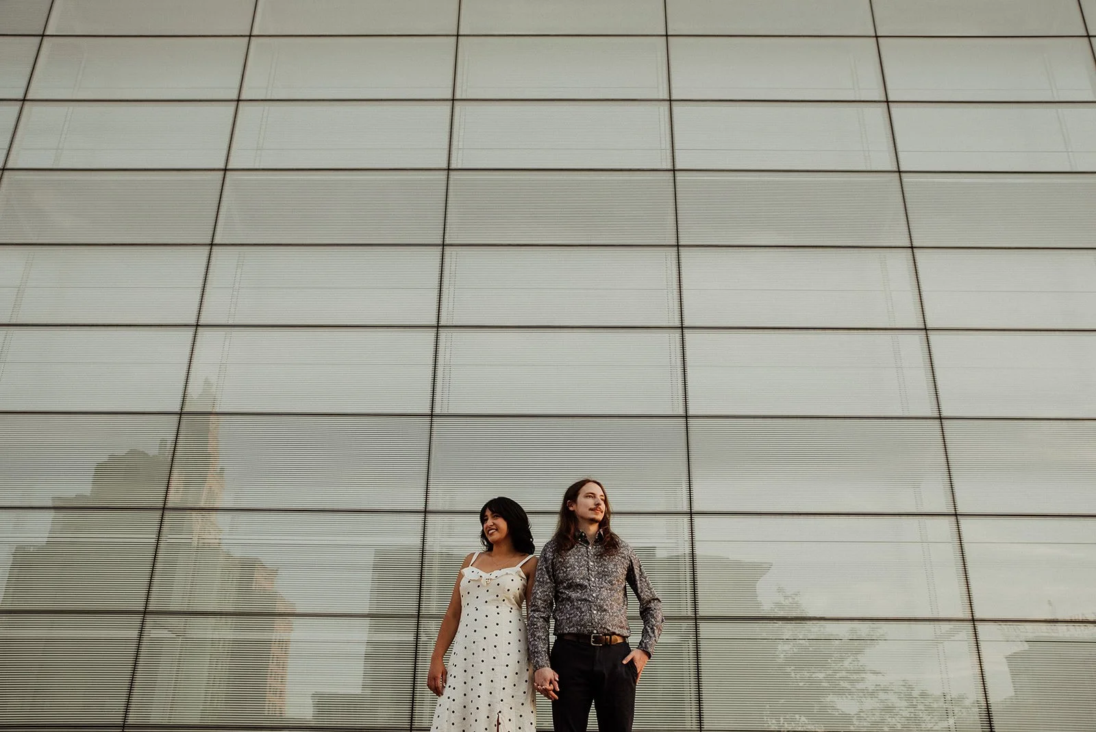 A man and woman standing outdoors in front of a reflective glass building, holding hands and smiling.