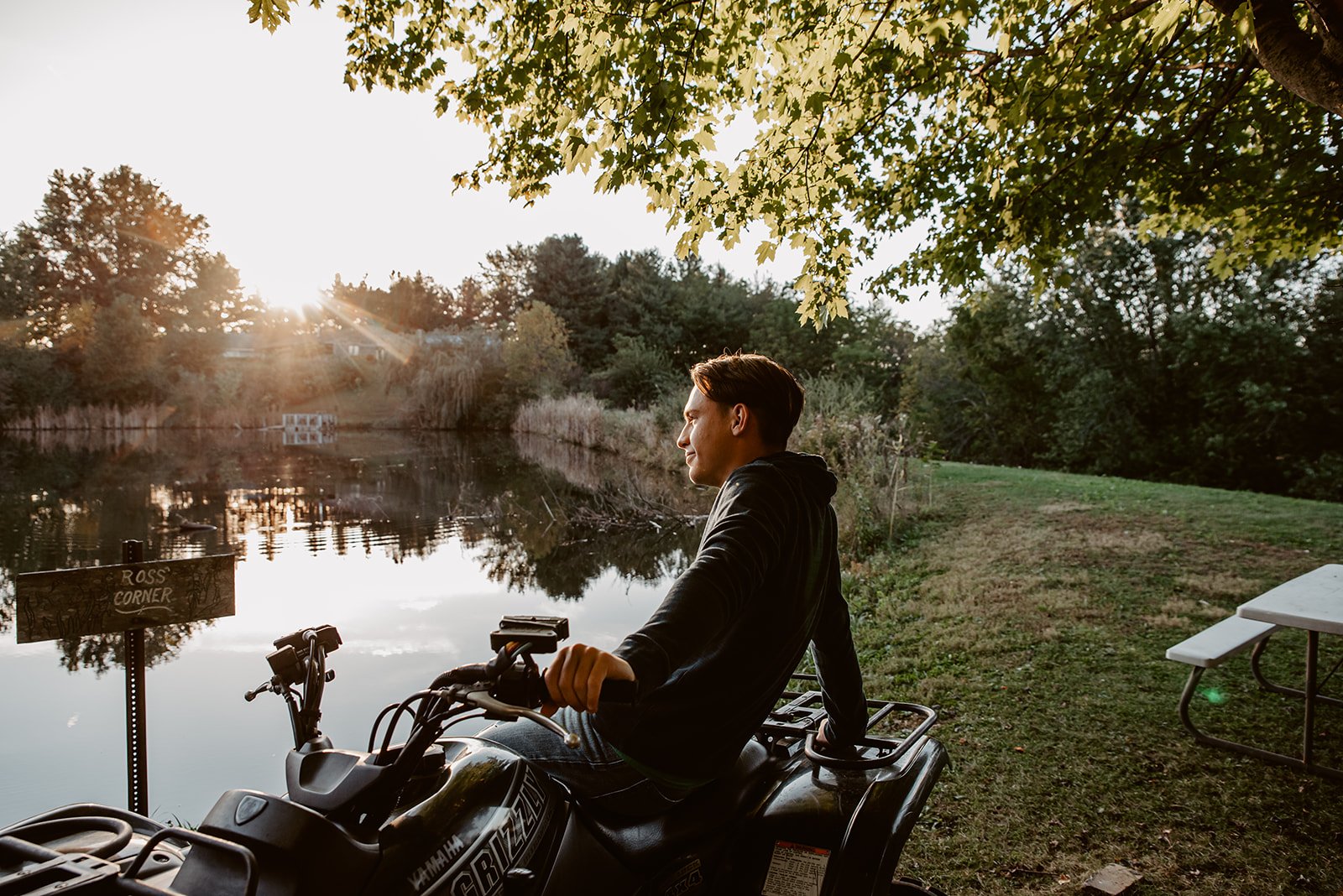 A young man sitting on a Yamaha all-terrain vehicle near a pond with trees and a setting sun in the background.