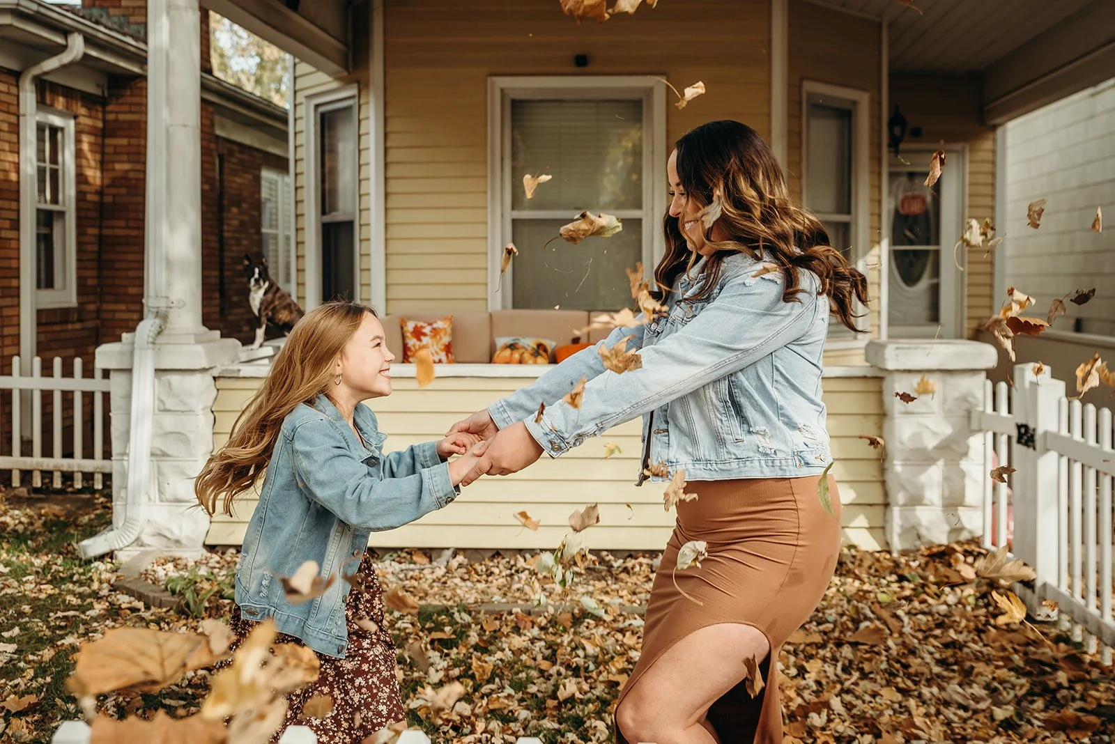 A woman and a young girl are playing together outside in front of a house, holding hands, surrounded by falling autumn leaves, with a cat sitting on the porch in the background.
