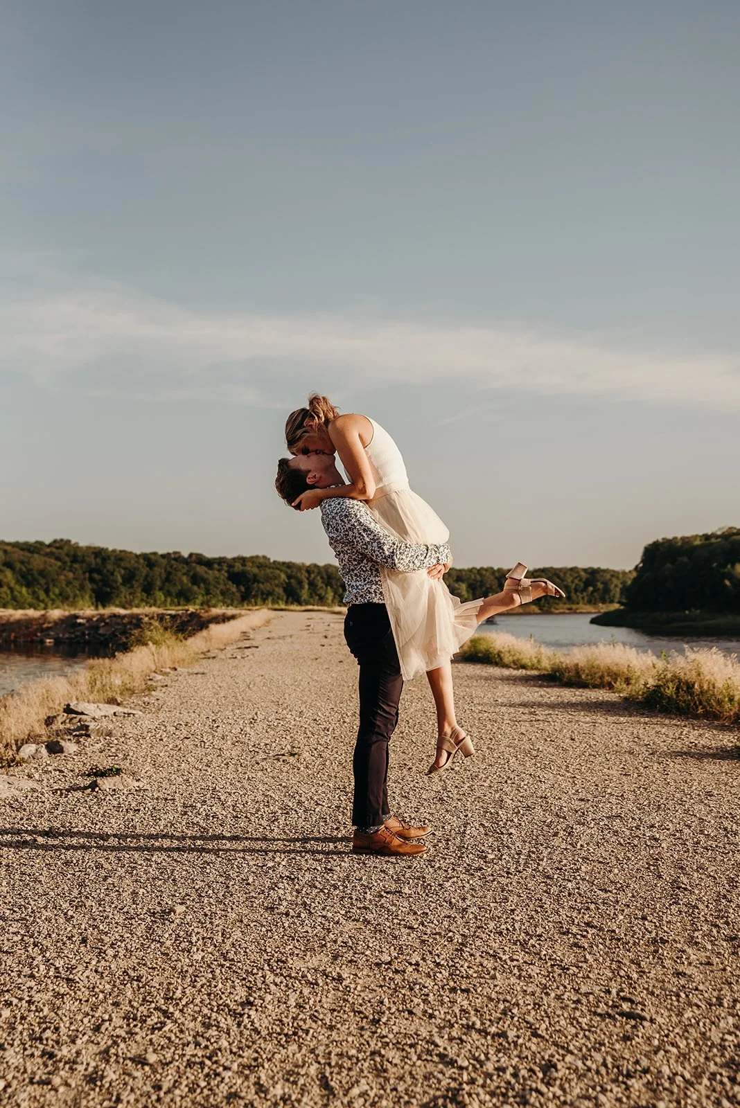 A couple dancing outdoors on a gravel path, with the man lifting the woman in his arms, near a river, with trees in the background under a clear sky.