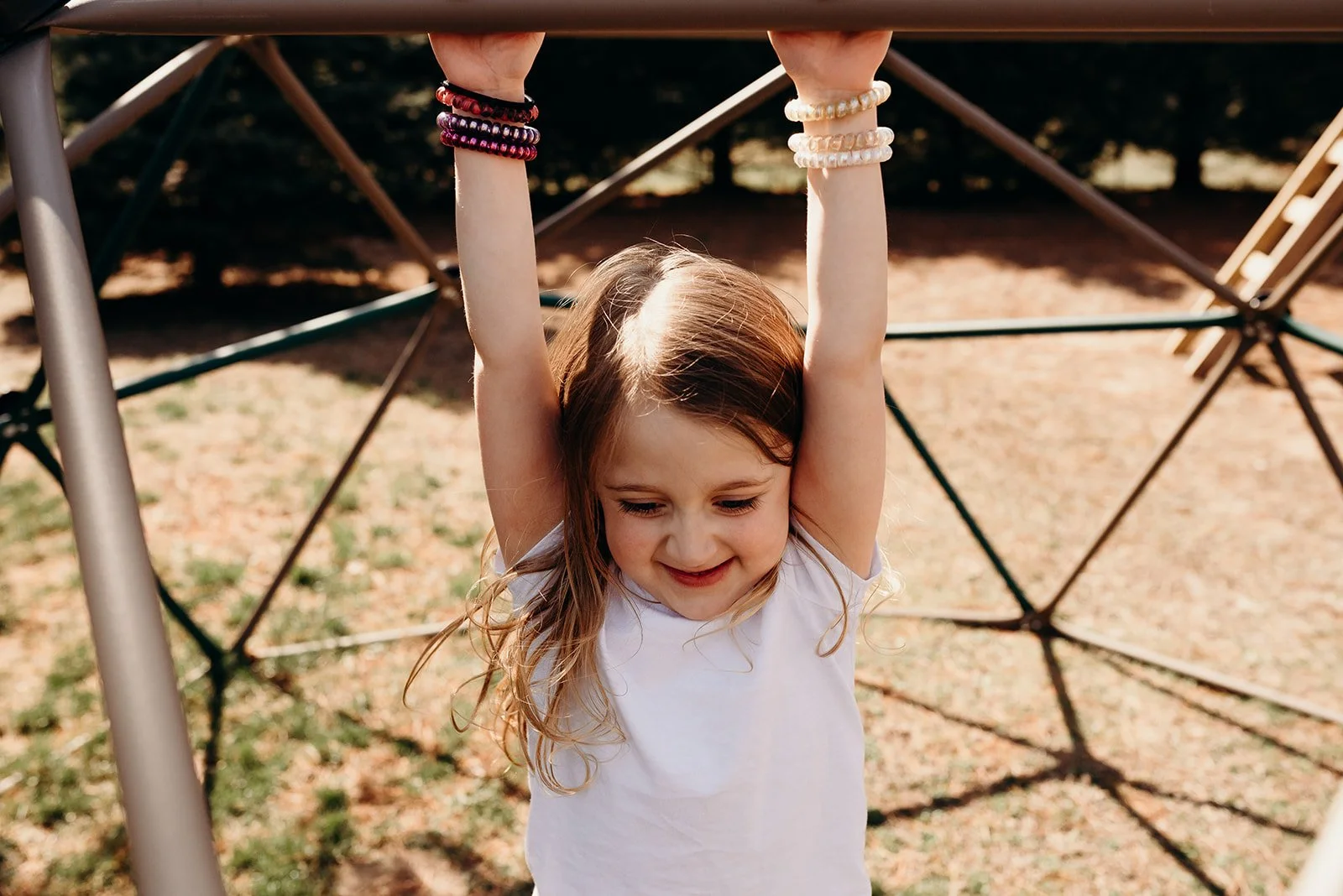 Young girl with long hair hanging down, hanging upside down from playground equipment, smiling with arms stretched upward, outdoors with ground covered in dirt and grass.
