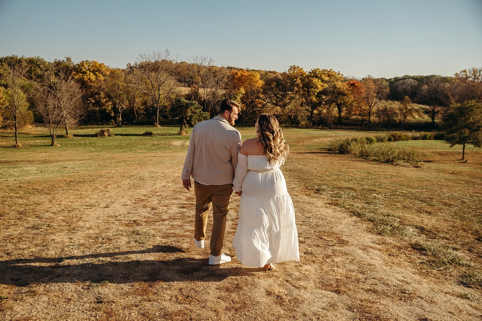 A couple walking hand in hand on a dirt trail in a park during autumn, with trees showing fall colors in the background.