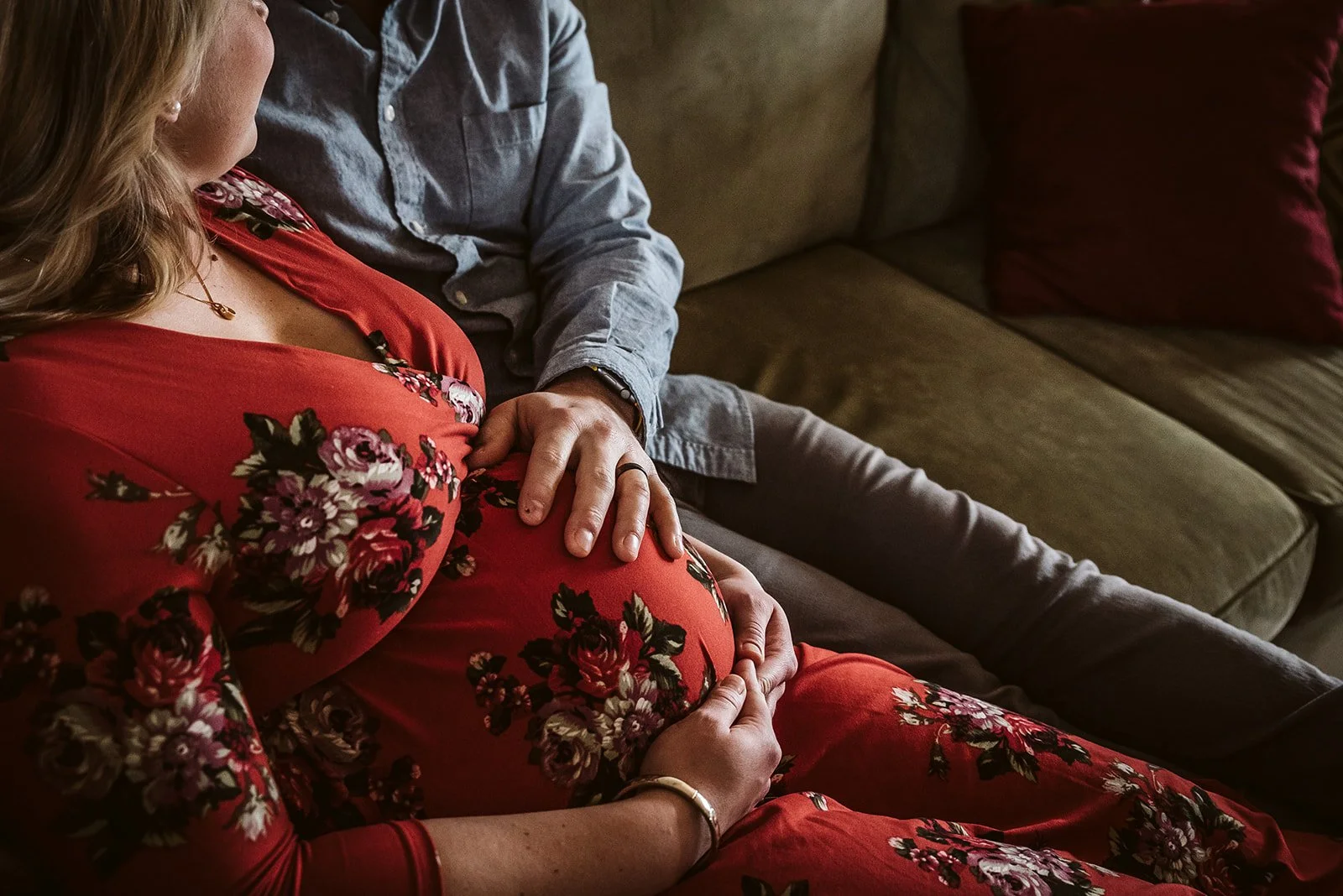 A pregnant woman in a red floral dress sitting on a sofa with a man in a blue shirt gently holding her pregnant belly.