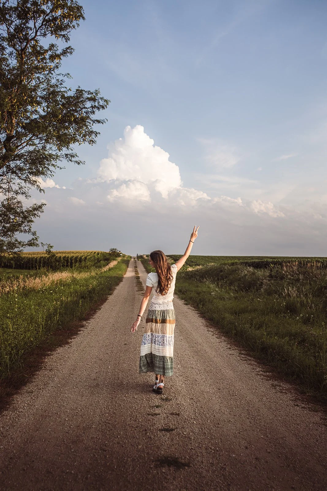 A woman walking down a dirt road in the countryside with fields on either side, raising one arm towards a partly cloudy sky.