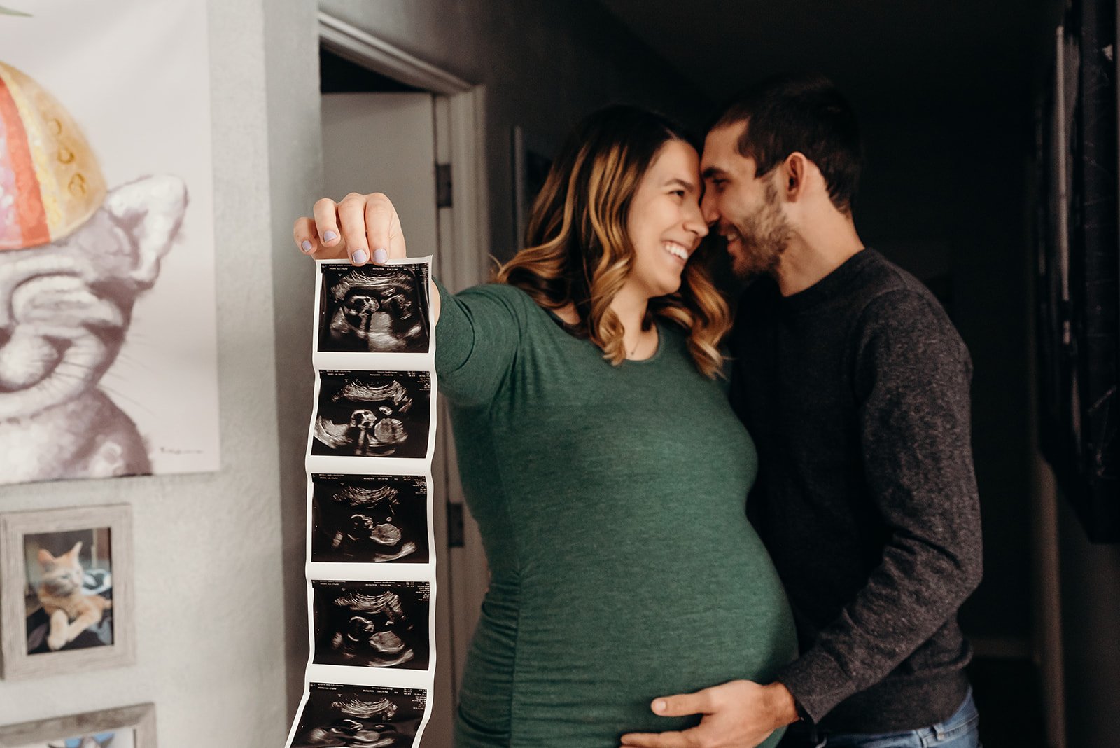 Pregnant woman and man touching foreheads, smiling, holding ultrasound pictures, in a home with wall art and a cat photo on the wall.