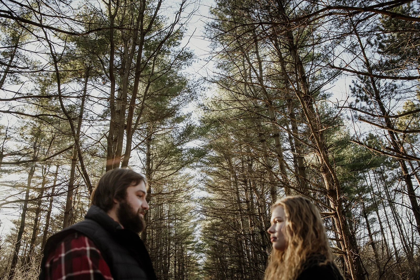 A man and woman standing close together in a forest with tall trees, looking at each other.