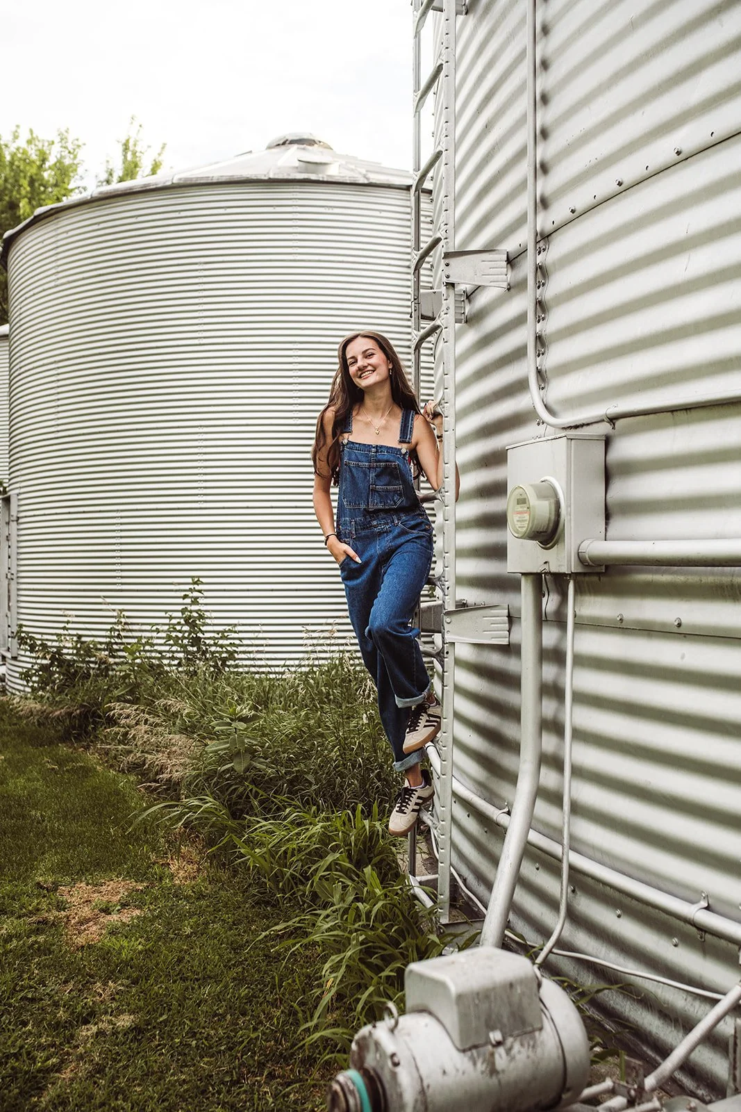 A young woman in a denim jumpsuit and sneakers smiling while climbing a ladder next to large metal grain silos outdoors.