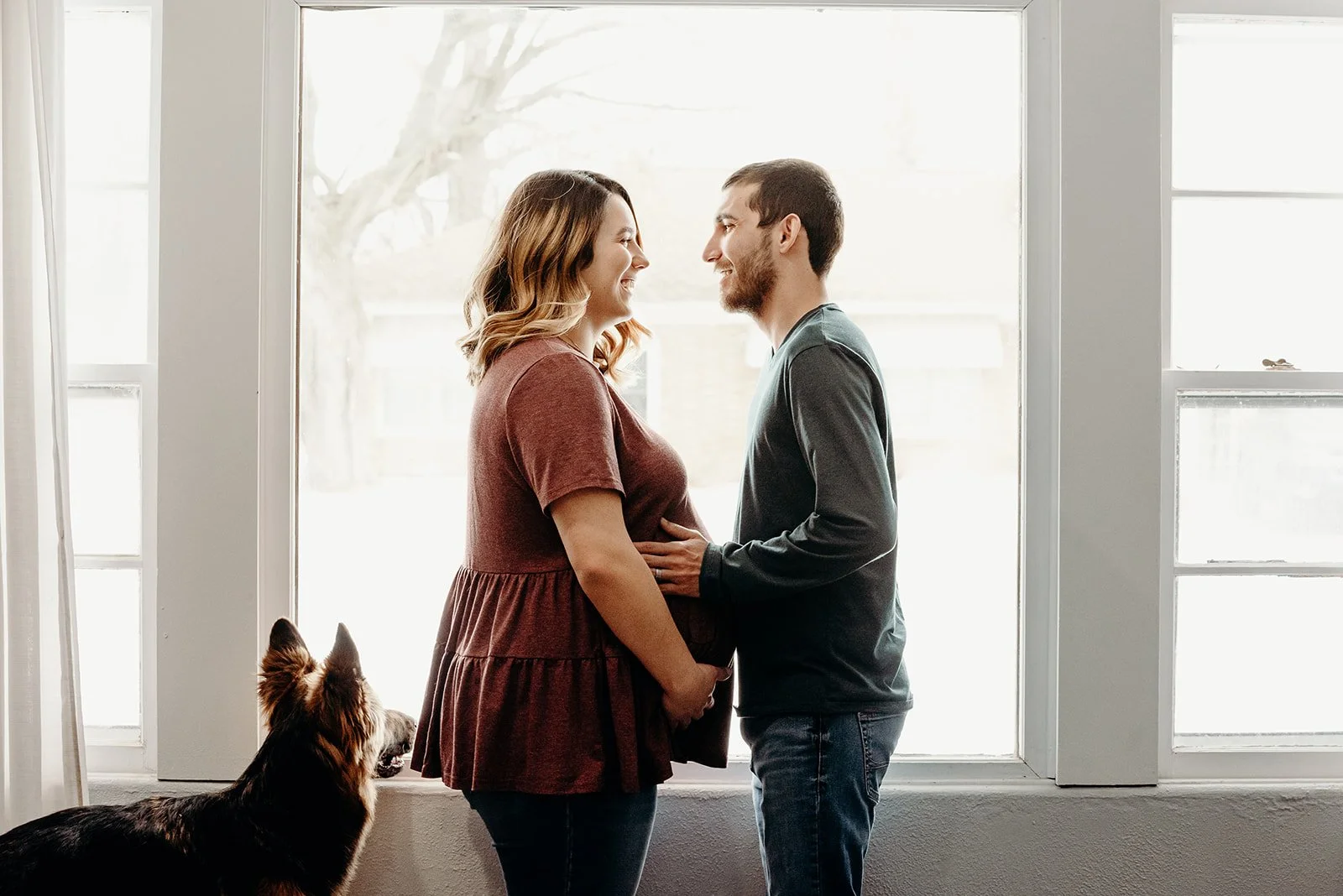 A couple stands facing each other in front of a large window, smiling and holding hands. A dog sits nearby, looking at the couple.