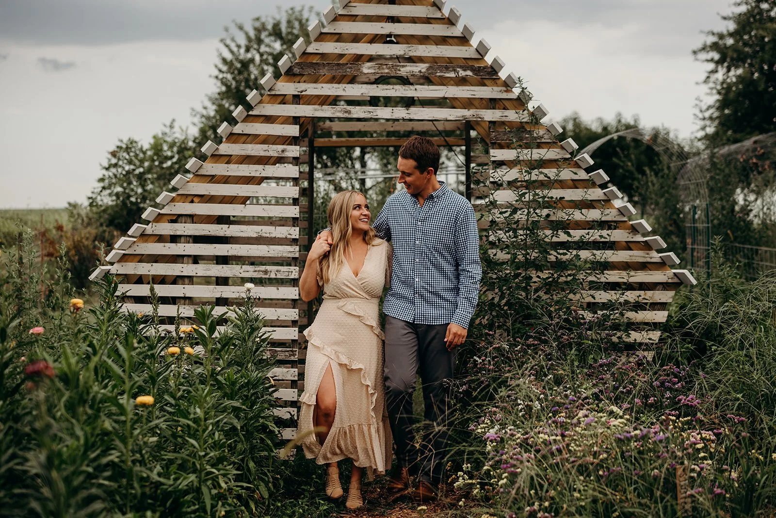 A couple walking through a garden, smiling at each other, standing in front of a wooden arched structure.