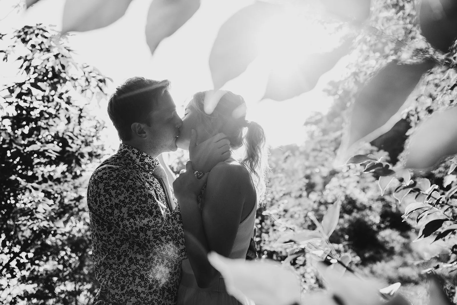A black and white photo of a couple sharing a kiss outdoors, surrounded by trees and sunlight filtering through the leaves.