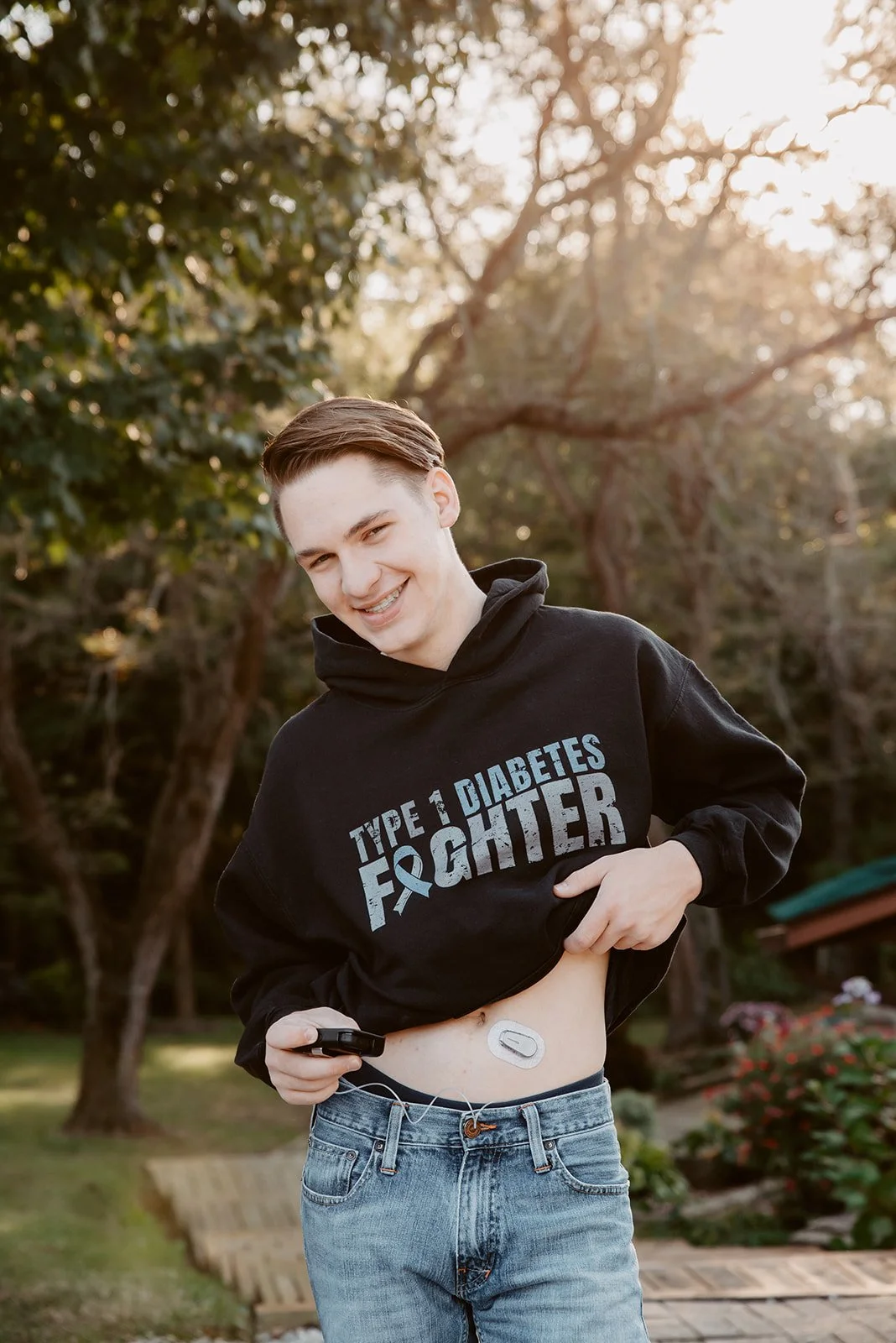 Young man with a smile lifting his black hoodie to reveal a diabetes insulin pump on his abdomen, outdoors in a park during sunset.