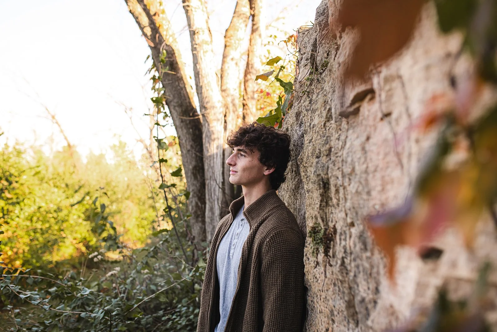 A young man with curly dark hair, wearing a gray shirt and brown jacket, leaning against a stone wall outdoors in a wooded area during autumn, with sunlight filtering through the trees.