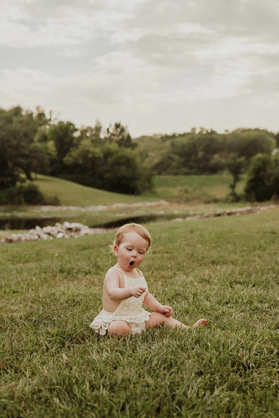 A young girl sitting on the grass near a body of water, with a surprised or excited expression on her face.