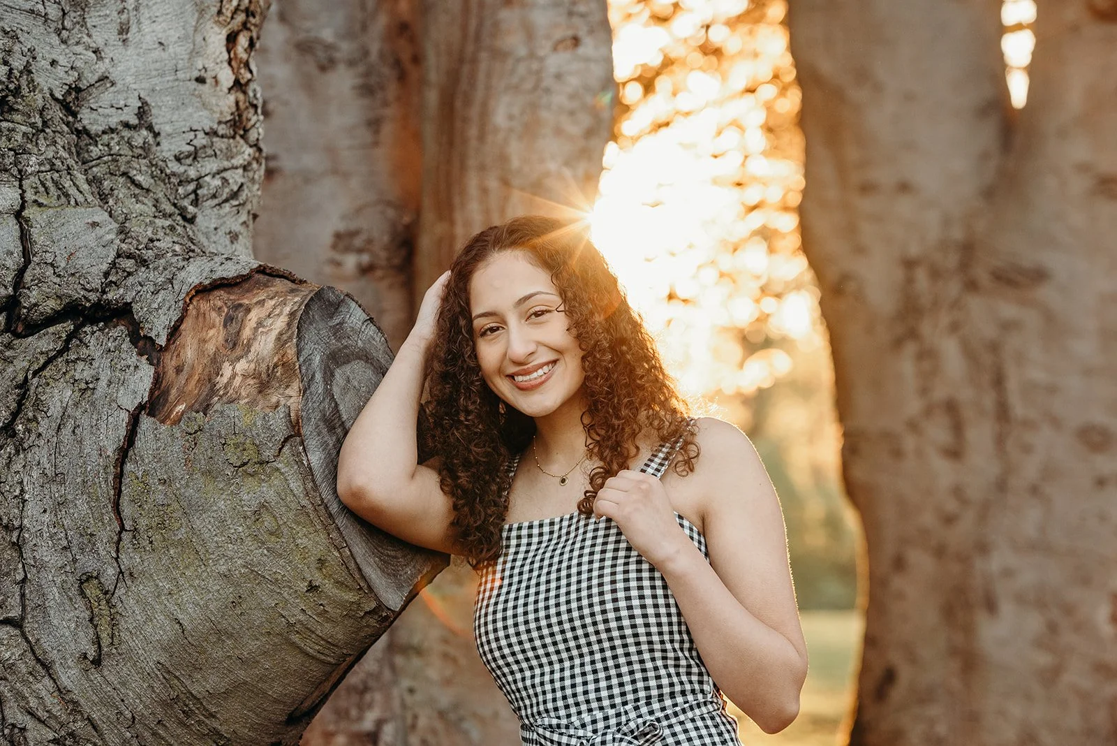 A young woman with curly hair smiling at the camera, standing outdoors near a large tree during sunset.