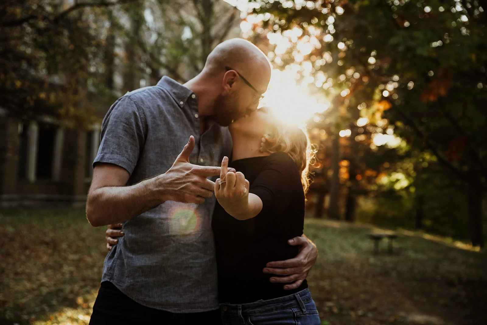 A couple sharing a kiss outdoors during sunset, with the man holding the woman around her waist and the woman holding his hand, with trees and sunlight in the background.