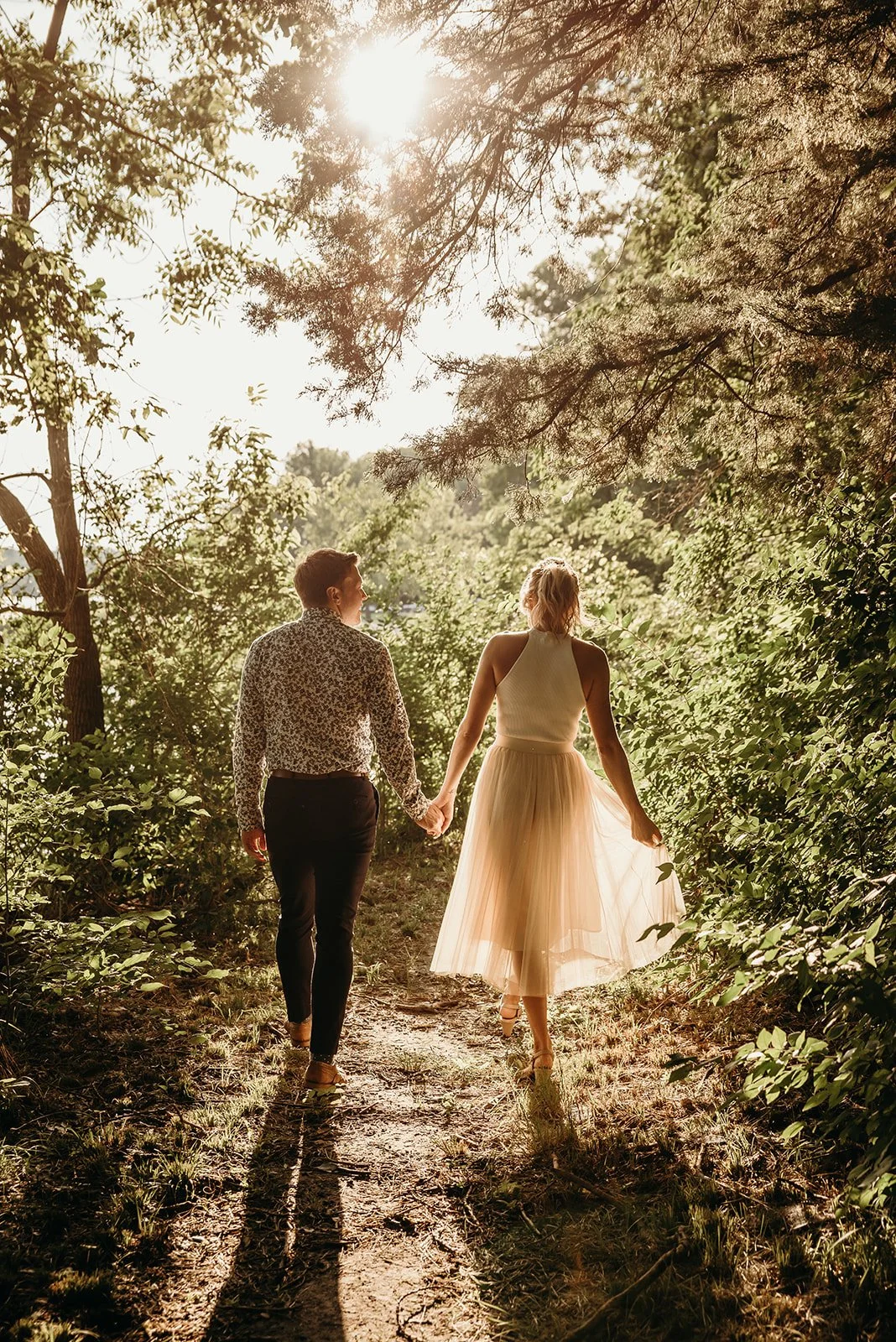 A couple holding hands walking on a forest trail during sunset, surrounded by lush green trees.