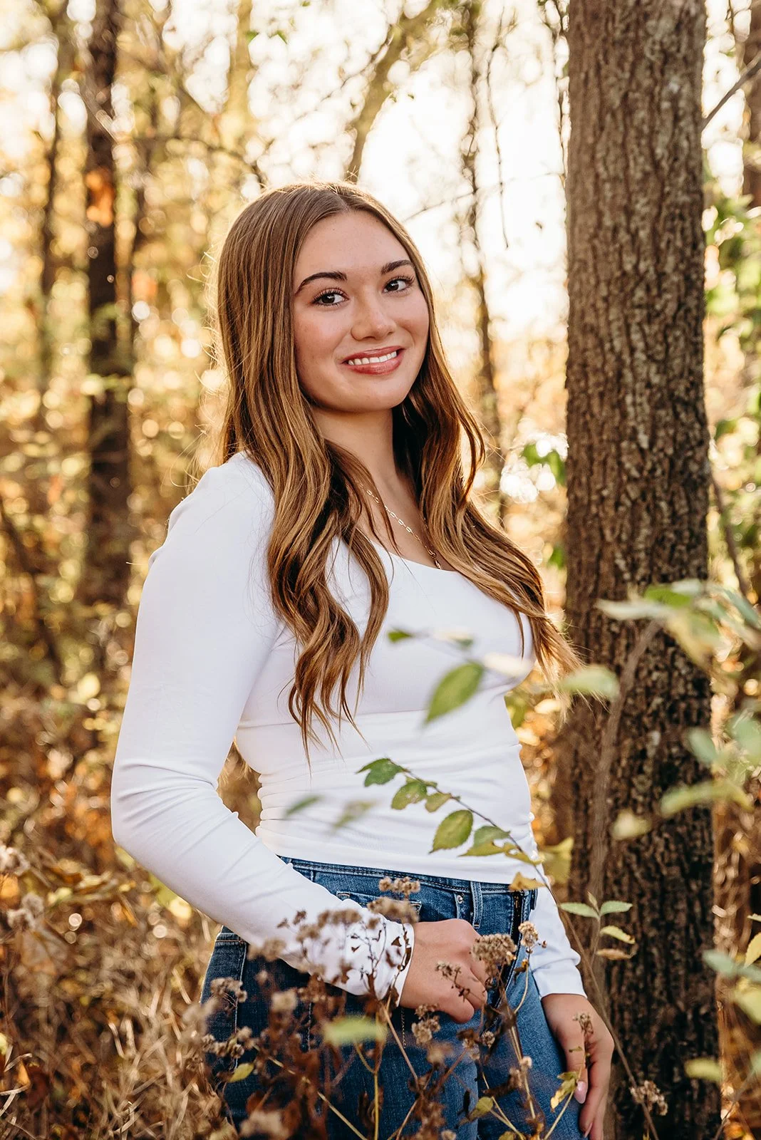 A young woman with long, wavy light brown hair standing outdoors among trees, wearing a white long-sleeve shirt and blue jeans, smiling at the camera during autumn.