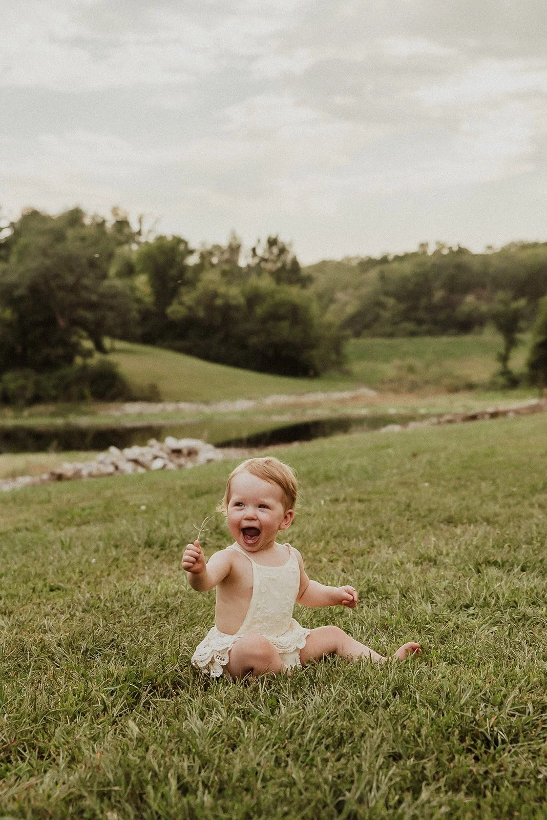 A happy toddler girl sitting on grass outdoors near a small river, holding a small stick, with green trees and a cloudy sky in the background.