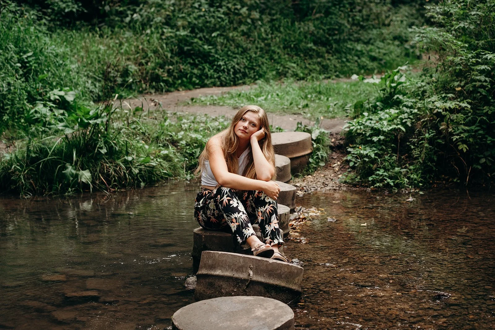 A woman sitting on stone steps in a creek surrounded by dense green foliage, resting her head on her hand and looking contemplative.