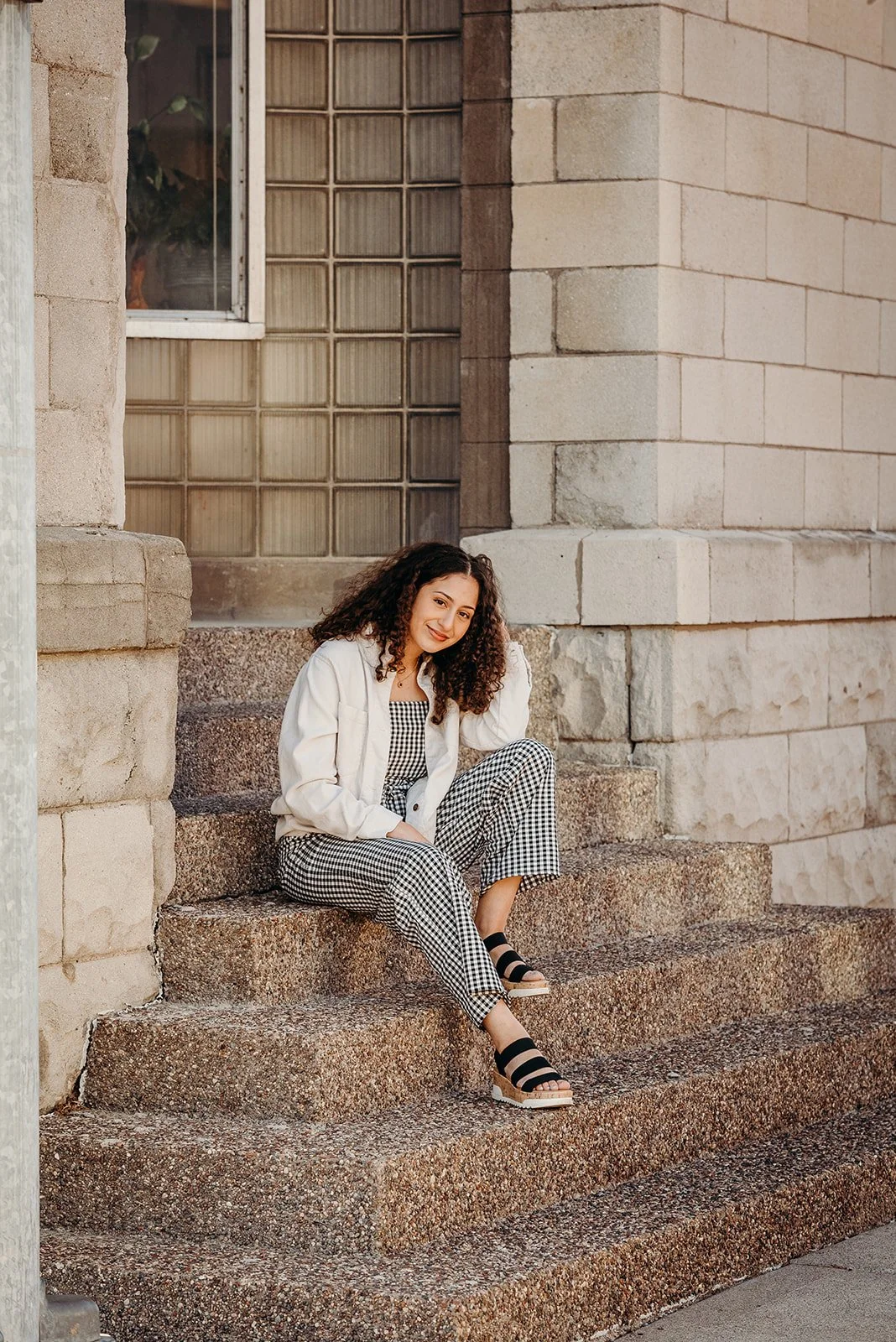 A woman with curly hair sitting on stone steps outside a building with a brick and glass block wall, wearing a white jacket, patterned pants, and black sandals, smiling at the camera.
