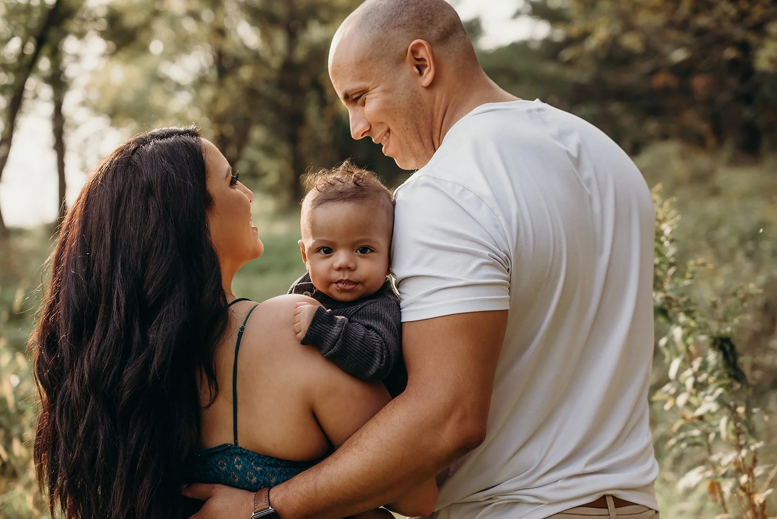 A happy family outdoors with the mother holding a baby and the father leaning in close, smiling at each other, during daytime.