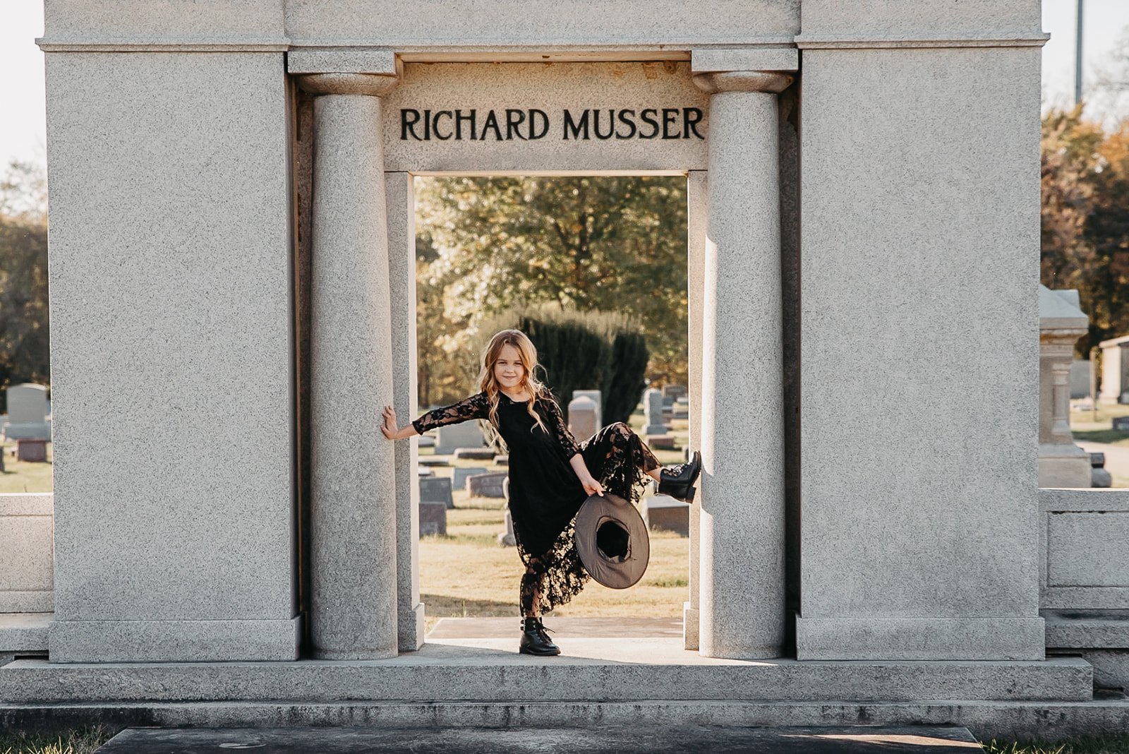A young girl in a black lace dress and black boots poses playfully at a cemetery monument with her hand on the stone column. She holds a wide-brimmed hat in her hand and is smiling. The monument has the name Richard Musser engraved at the top, and th