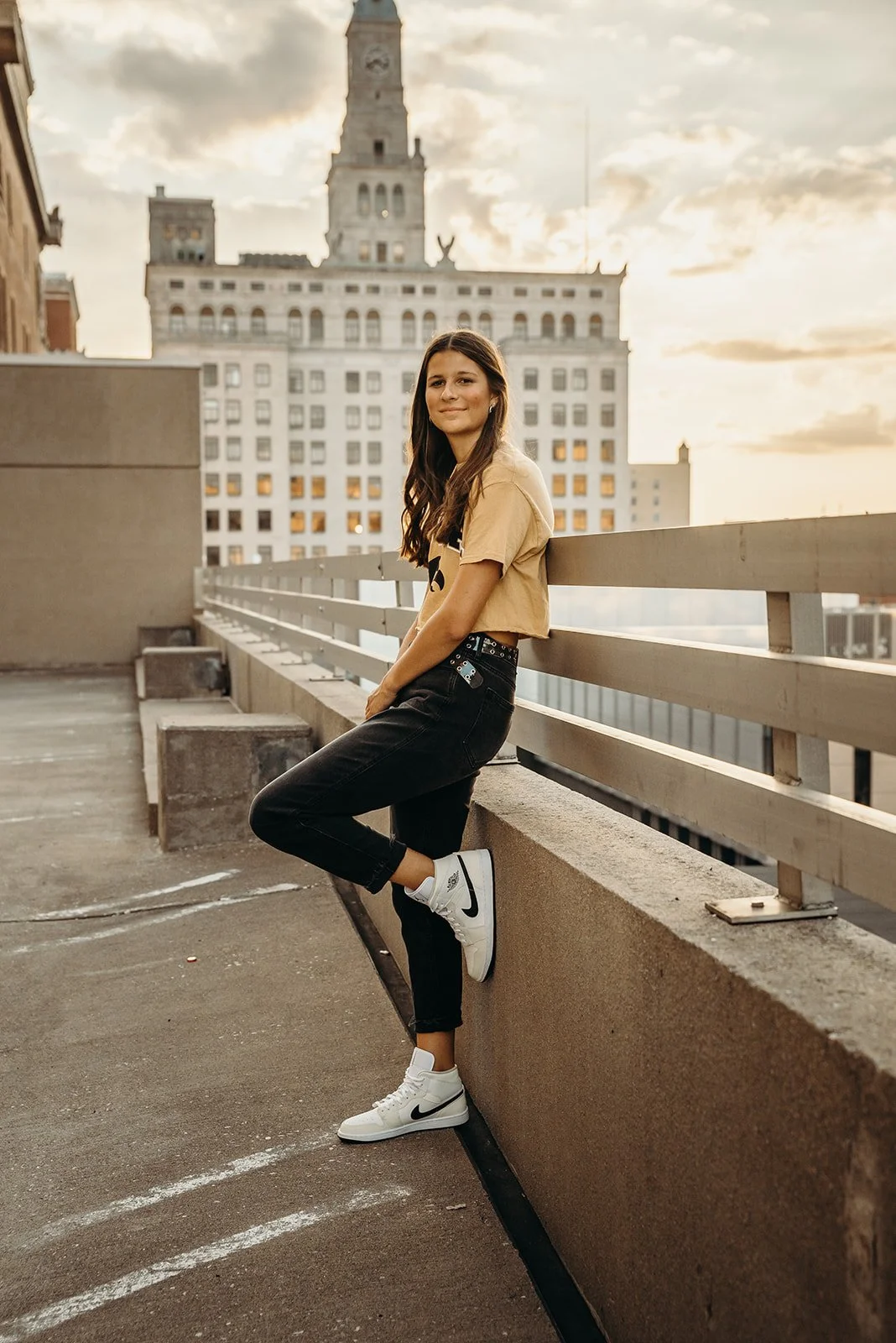 Young woman standing on a rooftop parking garage, leaning against the wall, with a city skyline and a tower in the background during sunset.