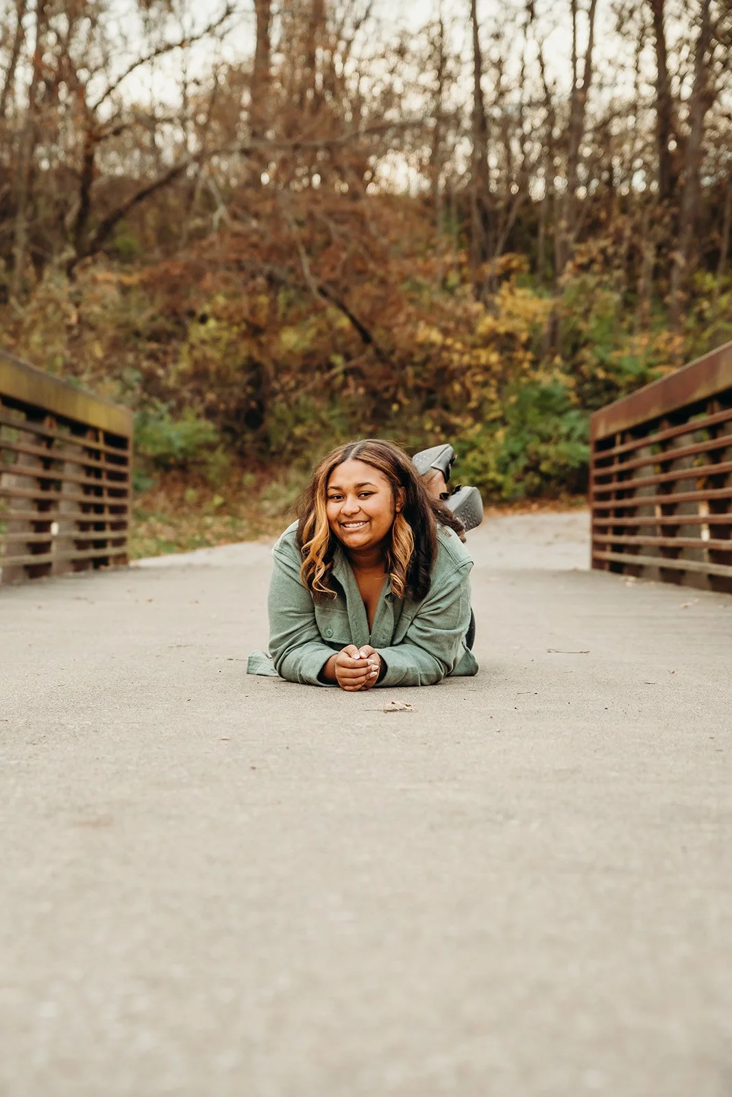 A woman lying on her stomach on a bridge with autumn trees in the background, smiling.