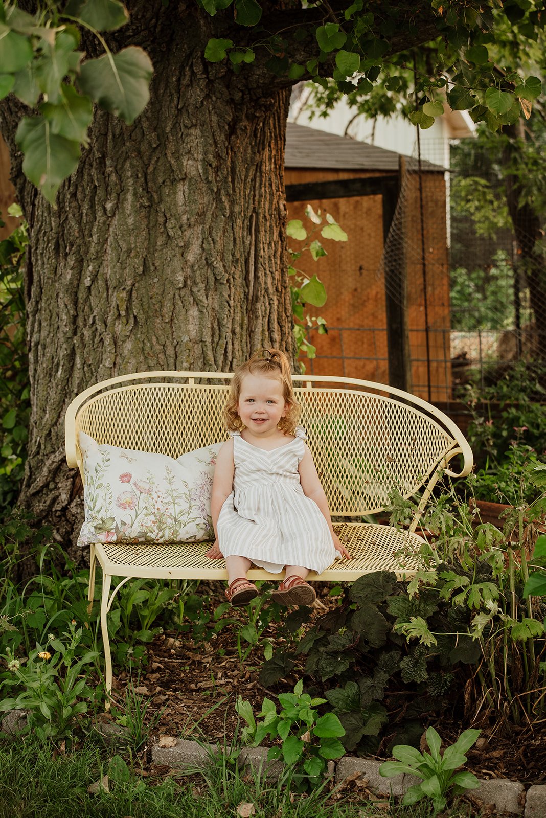 A young girl with curly red hair sitting on a yellow metal garden bench, smiling, in a garden with green plants and a large tree trunk behind her.