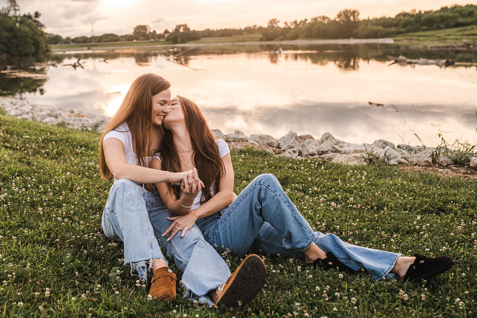 Two women sitting on grass near a lake during sunset, sharing a kiss and holding hands.