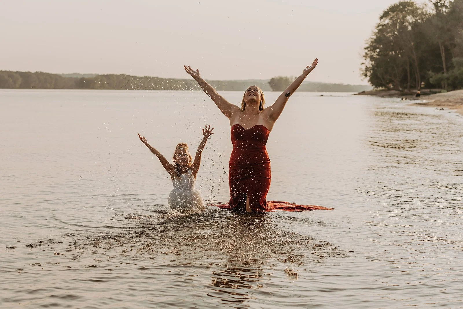 Woman and girl enjoying themselves in the water at a lake, with arms raised and water splashing around them, during sunset.