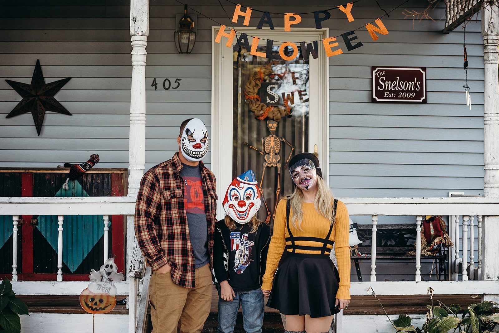Three people in Halloween costumes standing in front of a house decorated for Halloween, with a 'Happy Halloween' banner, a skeleton decoration in the doorway, and festive Halloween items, including a ghost sign and a pumpkin sign.