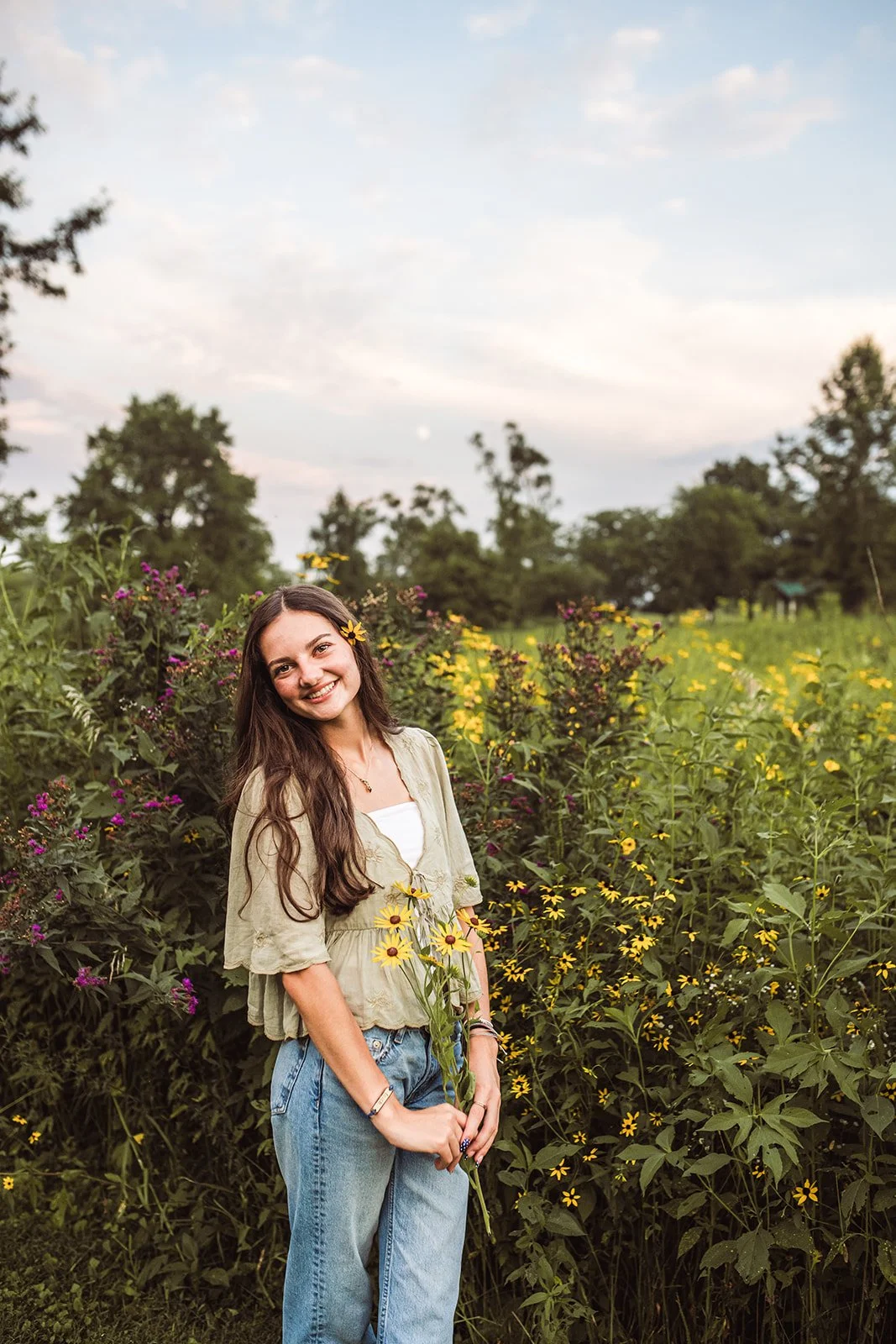 Smiling young woman with long hair holding yellow flowers, standing in a lush field of yellow and purple wildflowers during sunset.