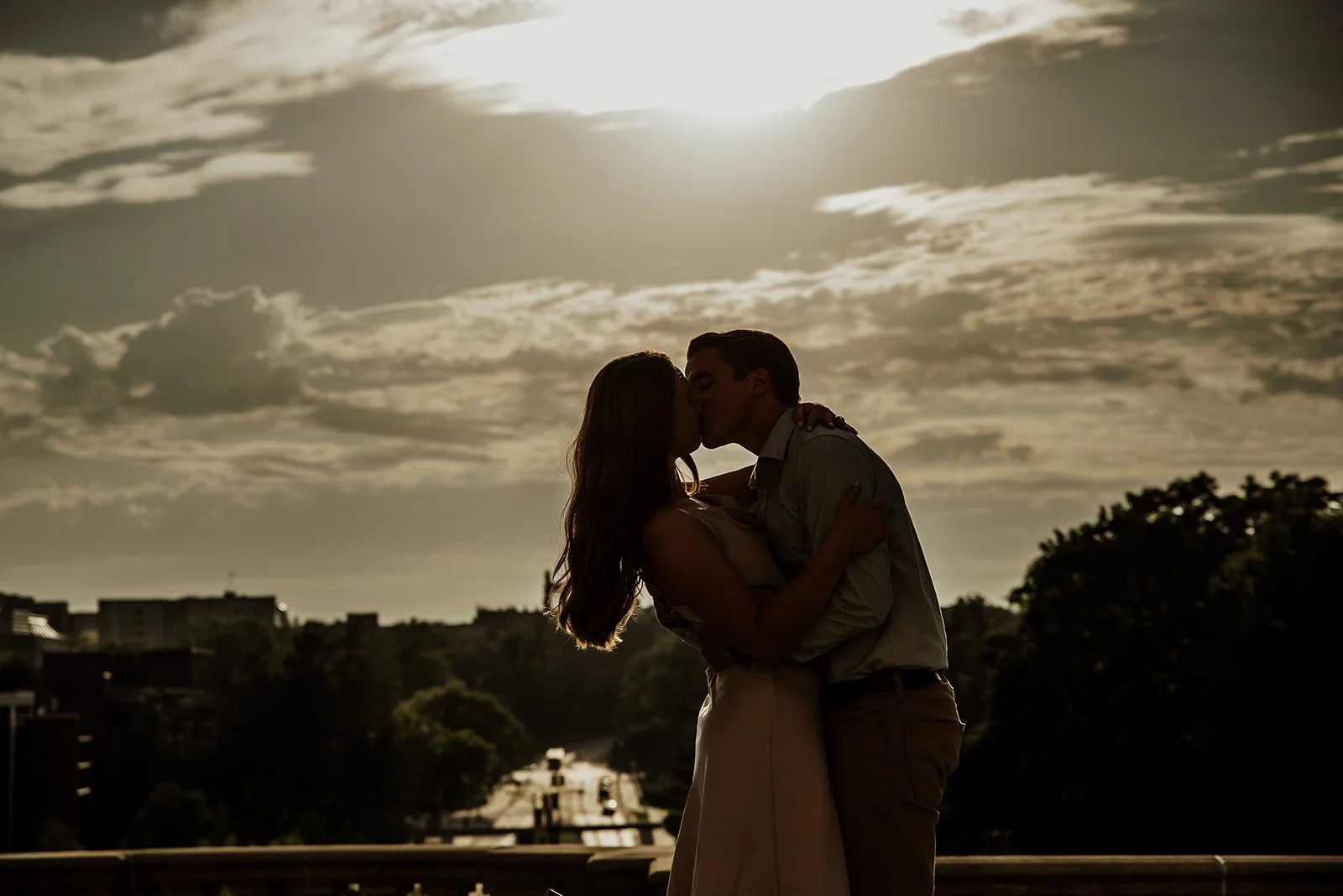 A silhouette of a couple kissing outdoors at sunset, with a sky filled with clouds and a cityscape in the background.