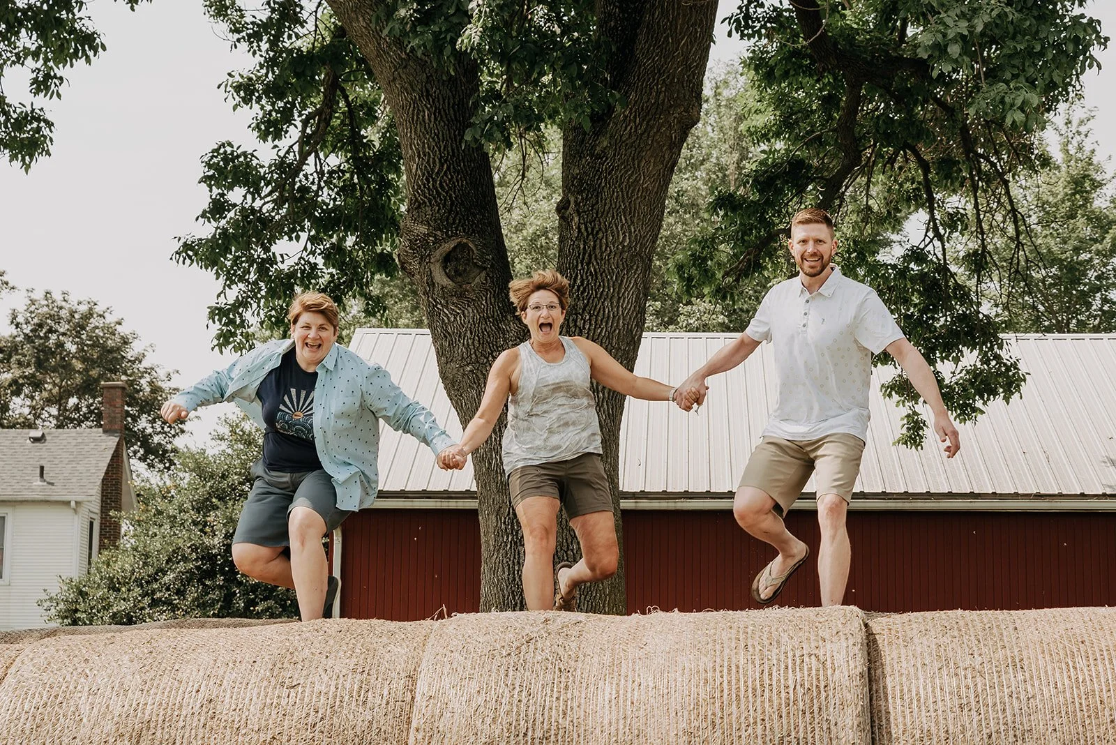 Three people holding hands and jumping on a hay bale outdoors, with a large tree and buildings in the background.