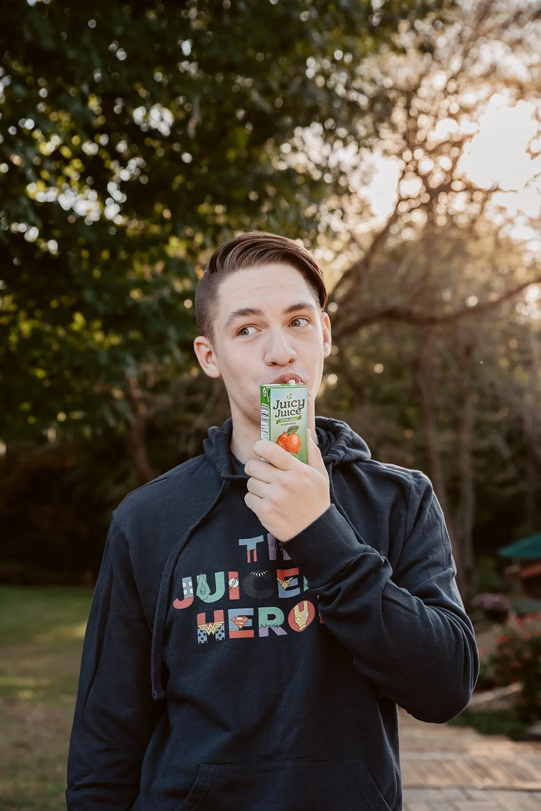 A young man with short hair wearing a dark hoodie with superhero logos and the phrase "Justice League" is holding a juice box near his mouth, outdoors with trees and sunlight in the background.